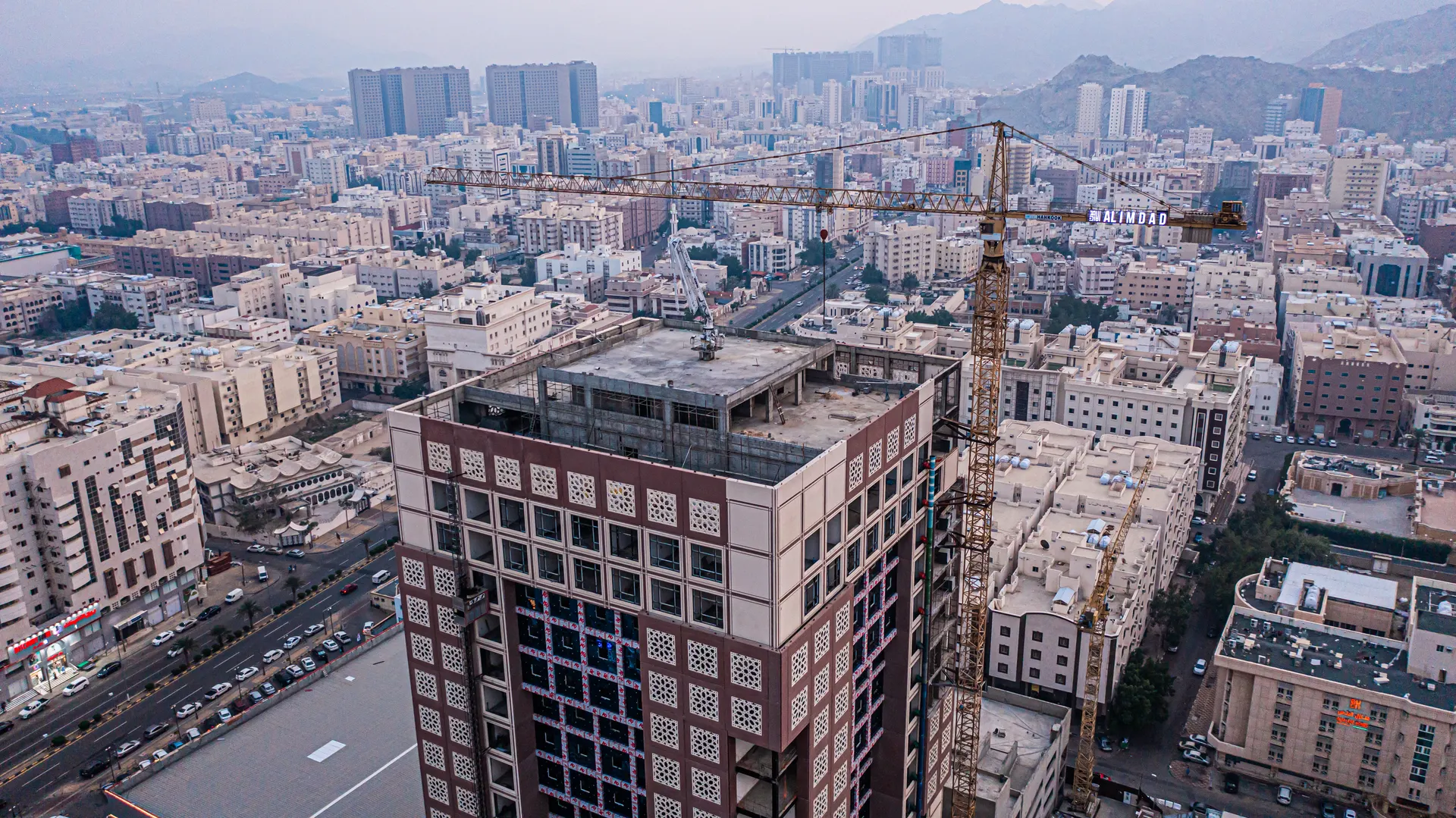 Aerial view of an Al Imdad tower under construction with a branded crane over the Makkah skyline at dusk.